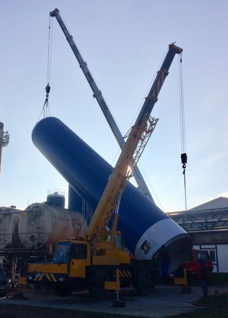 Dwa dźwigi marki NEKO podnoszą poziomo ustawiony, pomalowany na niebiesko-biało zbiornik przemysłowy na terenie zakładu produkcyjnego.