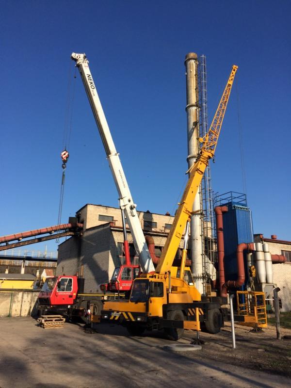 Dwa dźwigi samochodowe, jeden biały z napisem NEKO i jeden żółty, stoją na placu budowy z kominem w tle, pod błękitnym niebem.