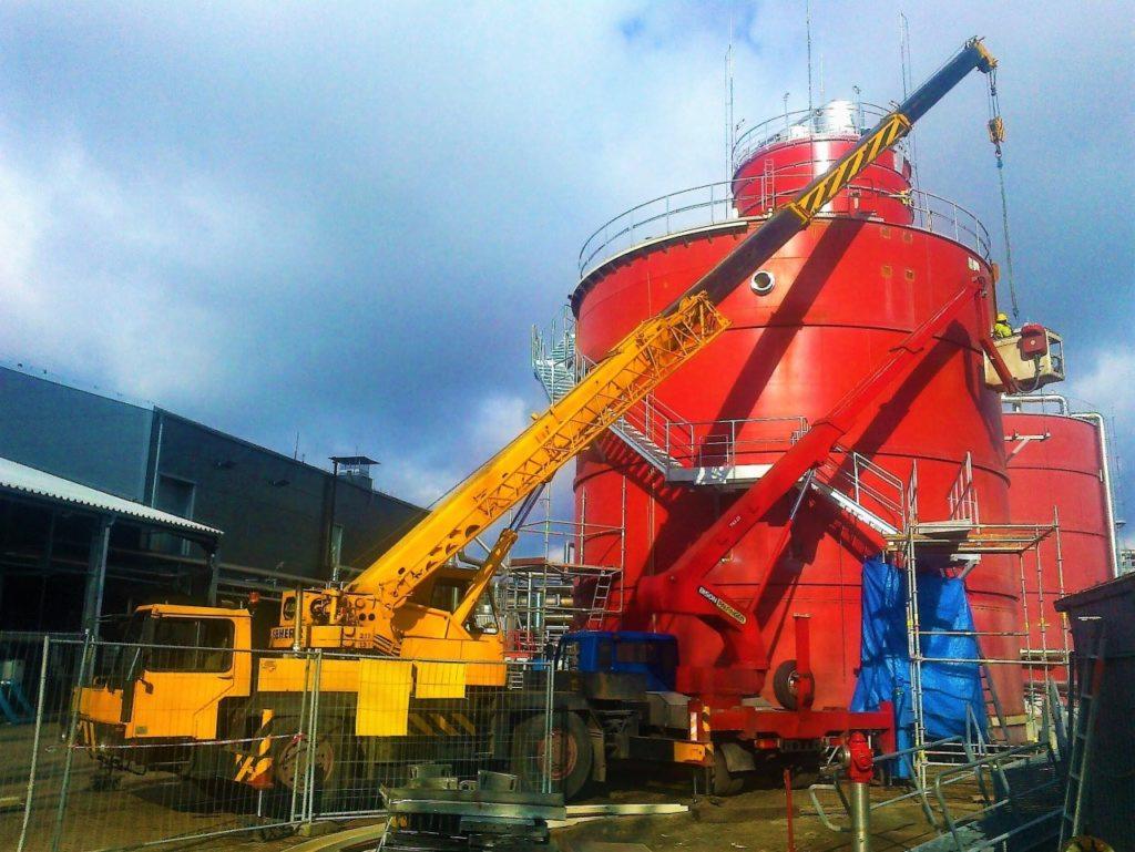 Żółty dźwig Liebherr i czerwony podnośnik koszowy Bison Palfinger pracują przy montażu lub konserwacji dużego, czerwonego zbiornika przemysłowego z metalowymi schodami i rusztowaniami, na tle...