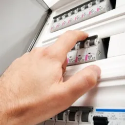 Otwarta skrzynka bezpiecznikowa z białymi wyłącznikami, dłoń przełącza jeden z nich w pozycji ON.