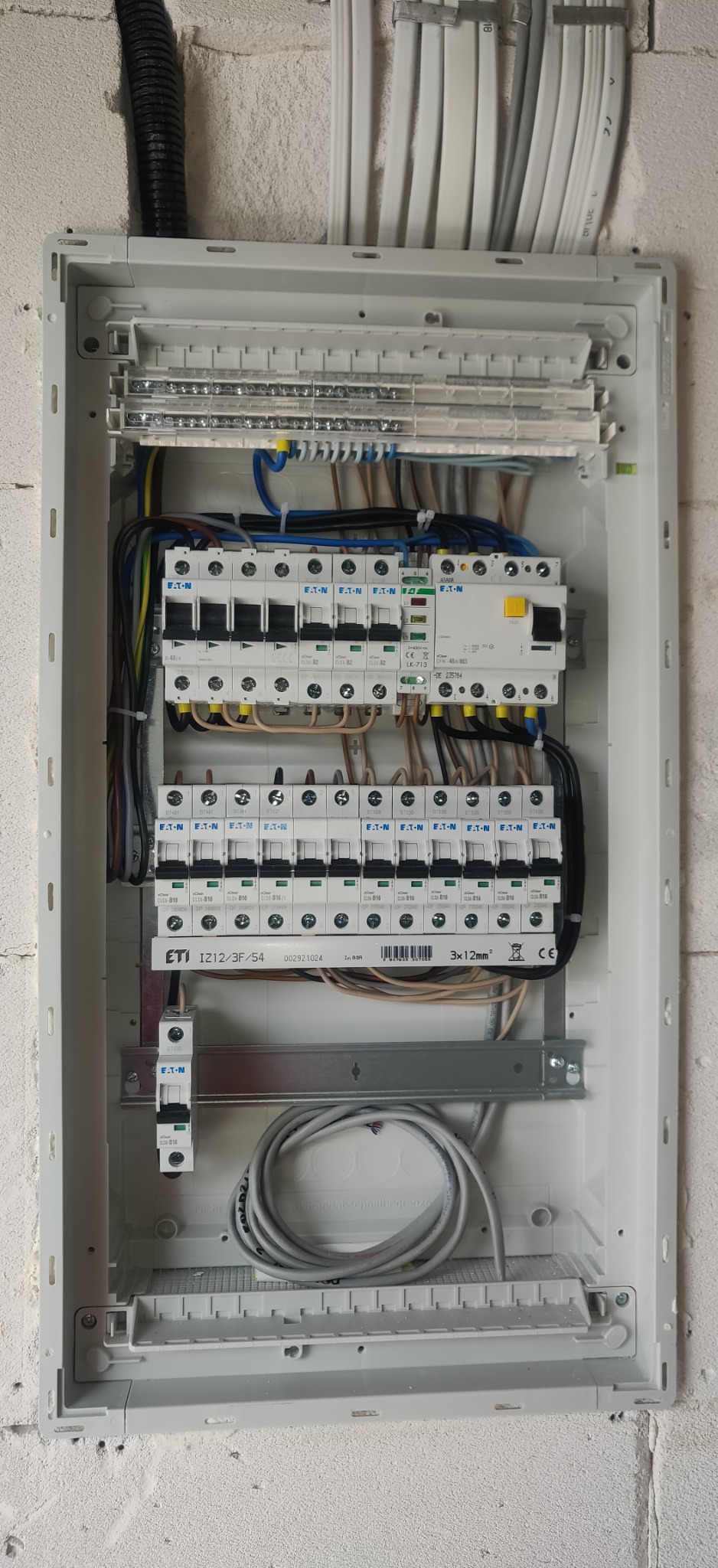 Otwarta skrzynka rozdzielcza z zamontowanymi bezpiecznikami ETI i Eaton, widoczne przewody i listwy zaciskowe, zamontowana na surowej ścianie z betonu komórkowego.