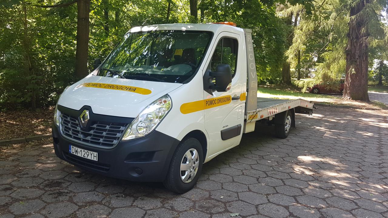 Biała laweta Renault Master z żółtymi pasami 'Pomoc Drogowa' na tle drzew w parku.