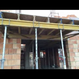 Wznoszenie stropu w budynku murowanym, widoczne podpory stalowe i szalunek drewniany, cegły silikatowe w ścianach, brak ocieplenia.