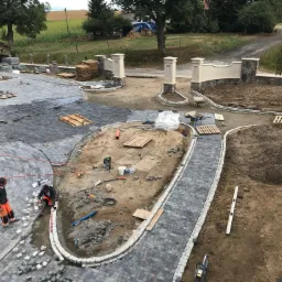 Układanie szarej kostki granitowej na posesji z widocznymi pracownikami w odblaskowych spodniach, narzędziami i częściowo ułożonym brukiem w tle krajobraz wiejski.