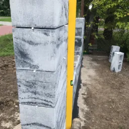 Wznoszony fragment ogrodzenia z szarych, cieniowanych bloczków betonowych, sprawdzany poziomicą, z widocznym gumowym młotkiem i elementami mocującymi.