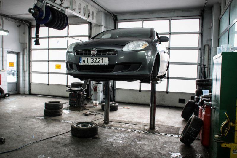 Szary Fiat Bravo na podnośniku w warsztacie samochodowym, widoczne opony, narzędzia i brama garażowa z oknami.