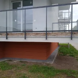 Szklana balustrada balkonowa z czarną ramą i kamienną okładziną poniżej, widok z dołu na elewację budynku z brązowym wykończeniem.