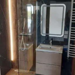 Nowoczesna łazienka z kabiną prysznicową typu walk-in, drewnopodobnymi płytkami i czarnym marmurem, oświetlonym lustrem nad umywalką z szafką.