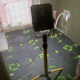 Monter - Podłoga w trakcie układania ciemnych płytek, widoczne krzyżyki dystansowe i lampa budowlana na trójnogu, fragment ściany z instalacją hydrauliczną.