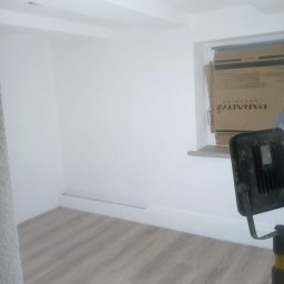 Monter - Wnętrze pokoju po remoncie, z jasnymi panelami podłogowymi, świeżo pomalowanymi białymi ścianami i sufitem, widoczna lampa budowlana na statywie oraz karton po płytkach ceramicznych przy oknie.