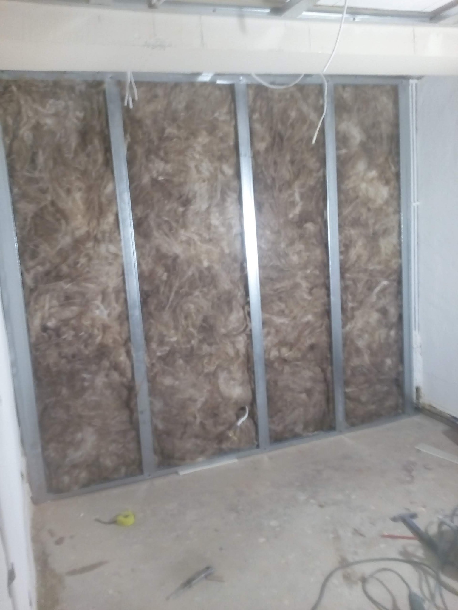 Ściana w trakcie izolacji wełną mineralną, widoczne metalowe profile i narzędzia na podłodze.