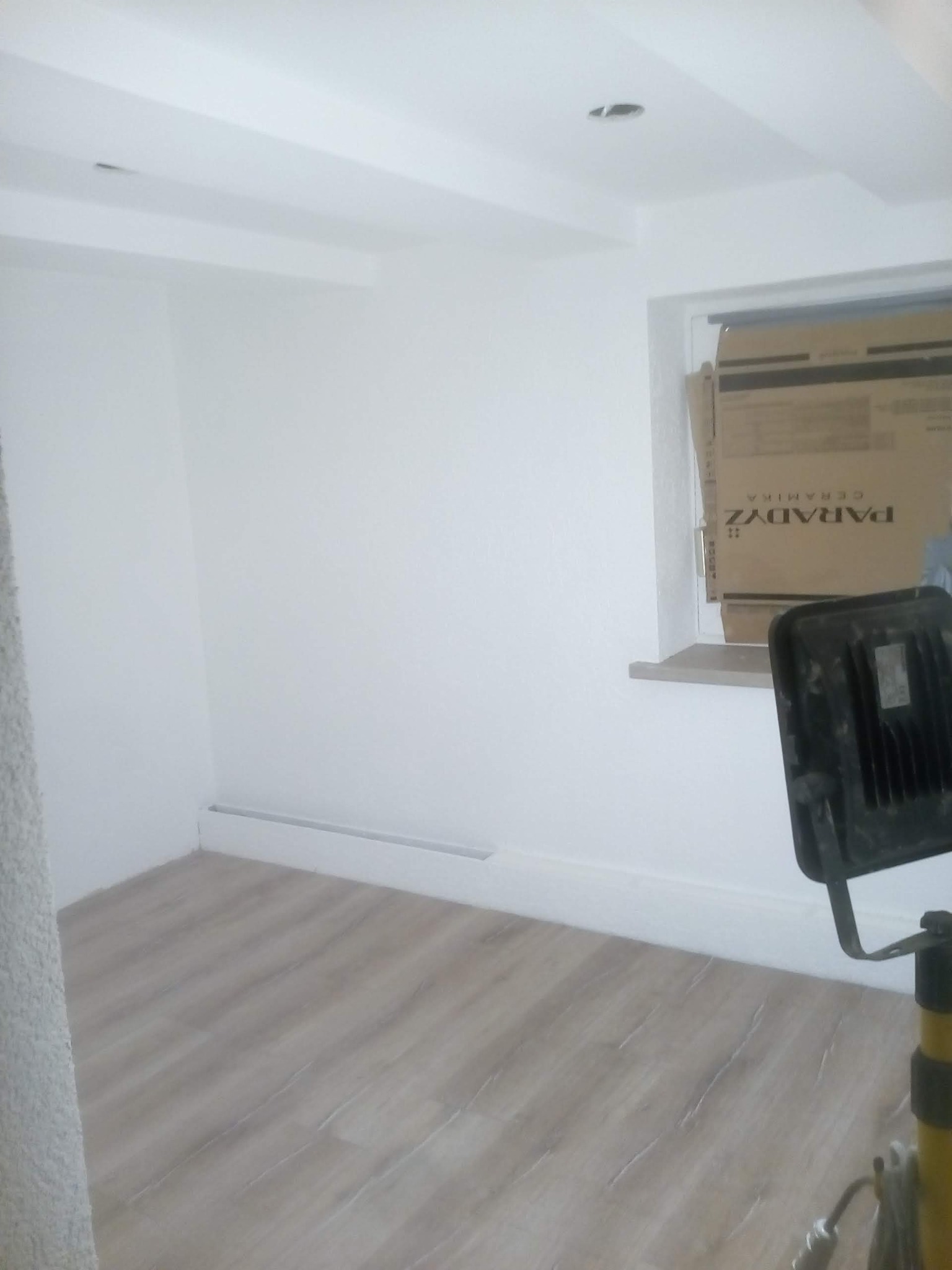 Wnętrze pokoju po remoncie, z jasnymi panelami podłogowymi, świeżo pomalowanymi białymi ścianami i sufitem, widoczna lampa budowlana na statywie oraz karton po płytkach ceramicznych przy oknie.