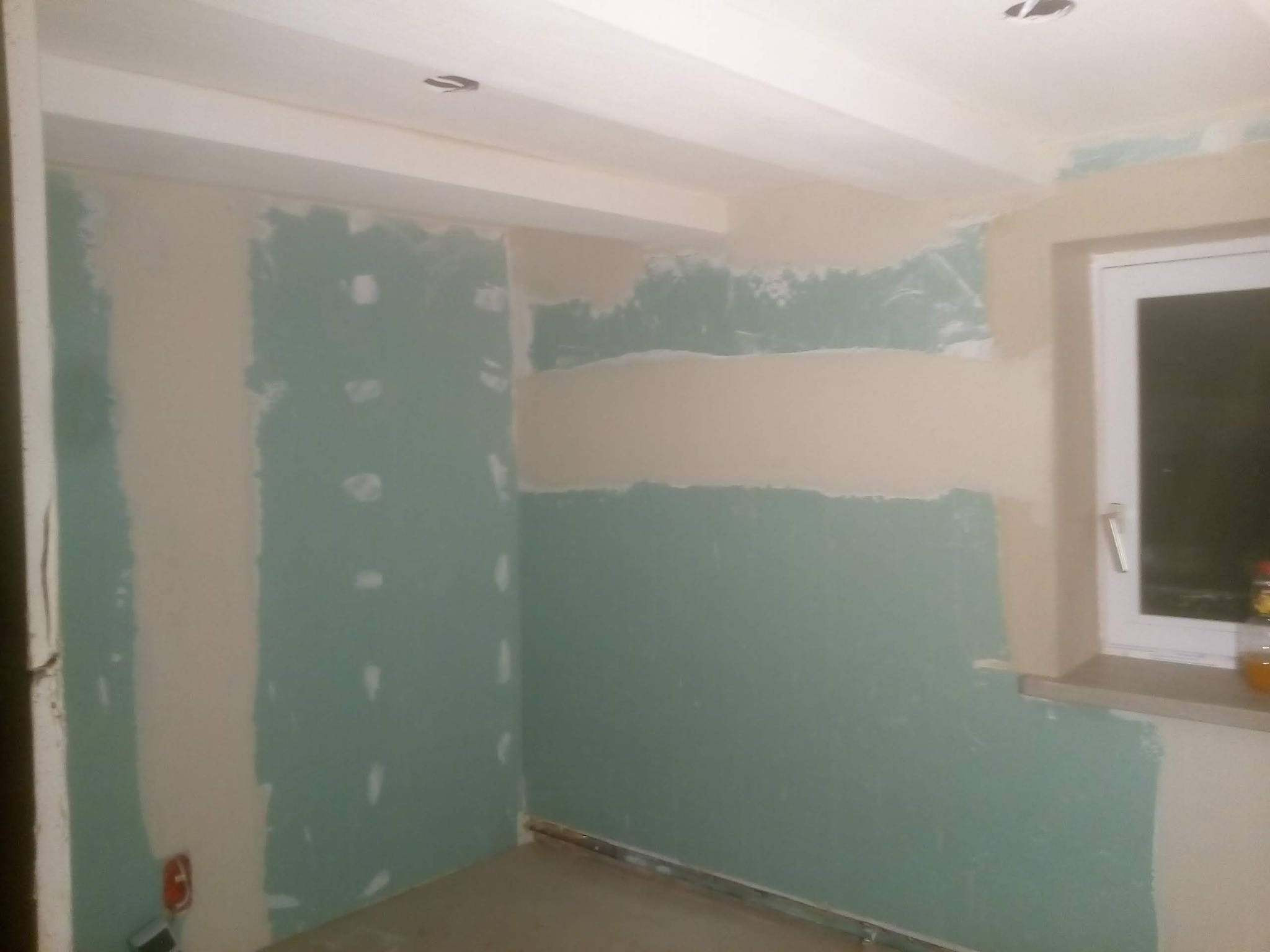 Pokój w trakcie remontu, ściany pokryte częściowo zieloną warstwą szpachli, widoczne łączenia płyt gipsowo-kartonowych, listwa przypodłogowa z metalu na podłodze, gniazdko elektryczne w dolnym...