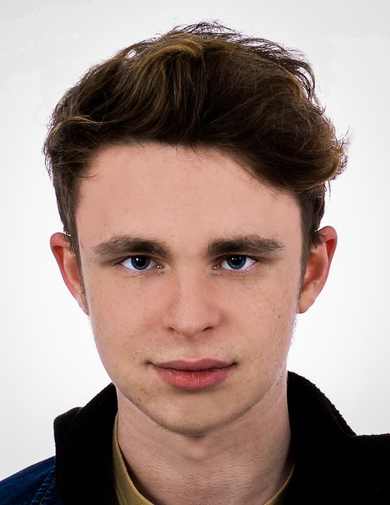 Portret młodego mężczyzny o jasnych oczach, z brązowymi włosami zaczesanymi do góry, na jasnym tle. Ubrany w ciemną kurtkę i koszulę w odcieniu musztardowym.