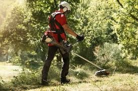 Mężczyzna w kasku i słuchawkach, z podkaszarką spalinową, kosi trawę na nierównym terenie z krzewami i drzewami w tle, w słoneczny dzień.