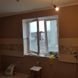 Wnętrze kuchni w trakcie remontu, z częściowo położonymi płytkami nad oknem, widoczne narzędzia na parapecie i surowa żarówka zwisająca z sufitu.