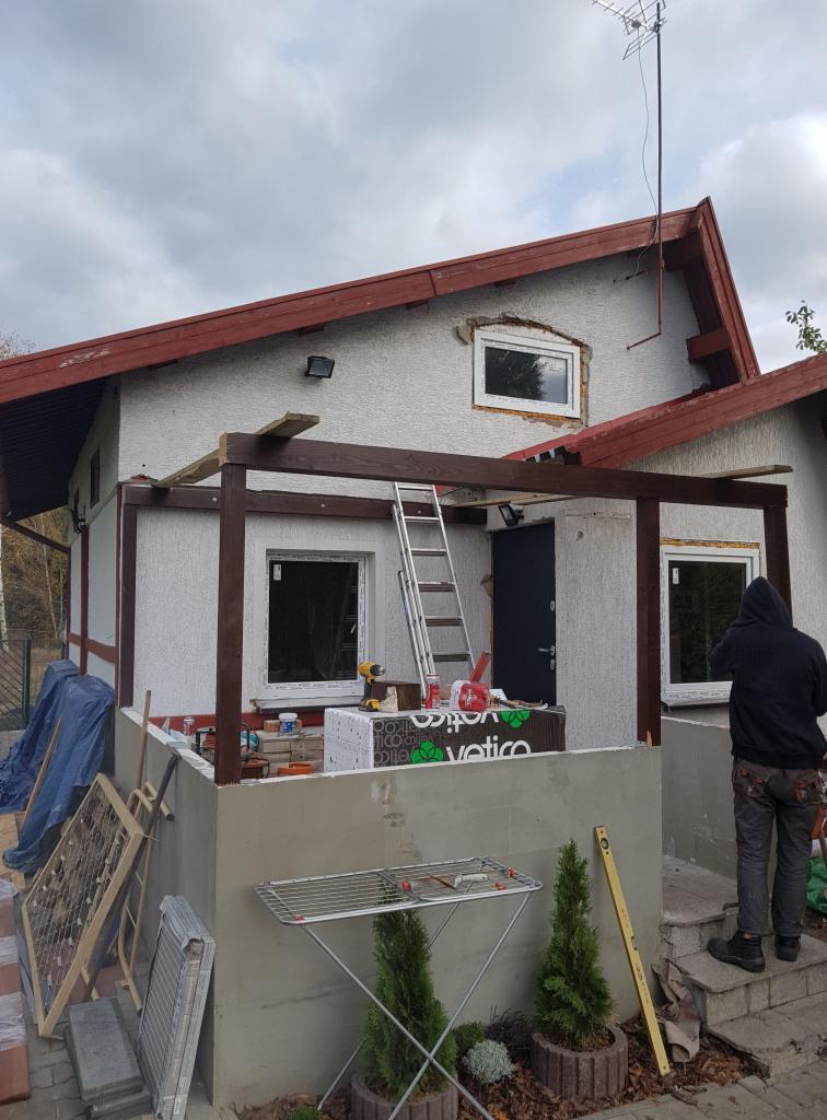 Budowa zadaszenia tarasu przy domu jednorodzinnym, widoczne drewniane belki konstrukcyjne, drabina oparta o ścianę, materiały budowlane i pracownik w bluzie z kapturem.