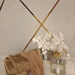 Złota, pikowana torba podróżna i mała kopertówka z weluru, leżące na białym futerku obok beżowej, połyskującej tkaniny, dekoracyjnych lampionów ze świecami i wazonu z białymi gałązkami, na tle...