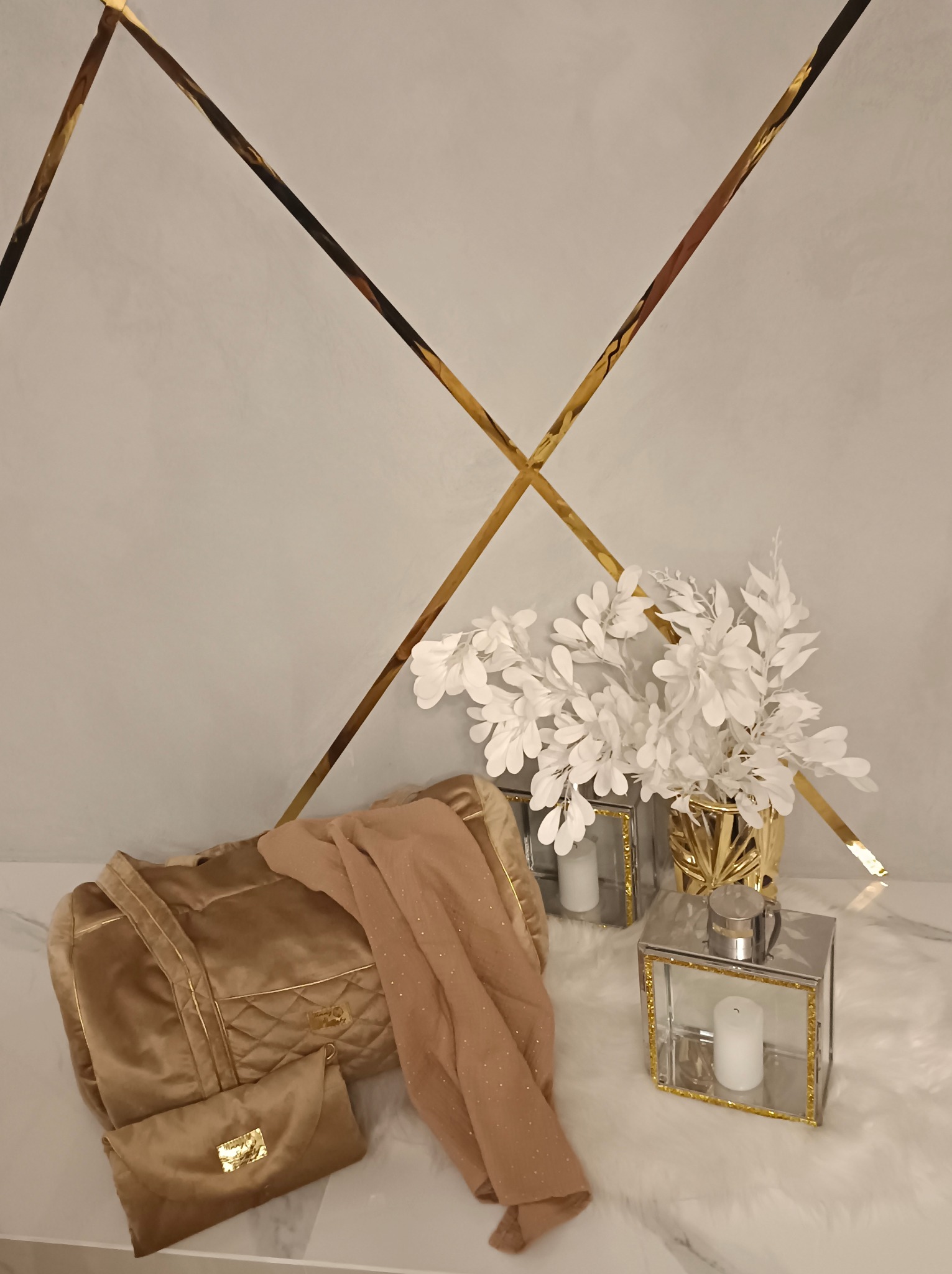 Złota, pikowana torba podróżna i mała kopertówka z weluru, leżące na białym futerku obok beżowej, połyskującej tkaniny, dekoracyjnych lampionów ze świecami i wazonu z białymi gałązkami, na tle...