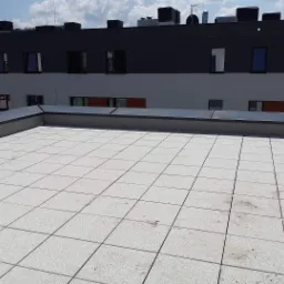 Wykończony taras na dachu budynku w Kadzidle, pokryty jasnoszarą, kwadratową płytką z delikatnym wzorem, z widocznym fragmentem elewacji i niebem w tle.