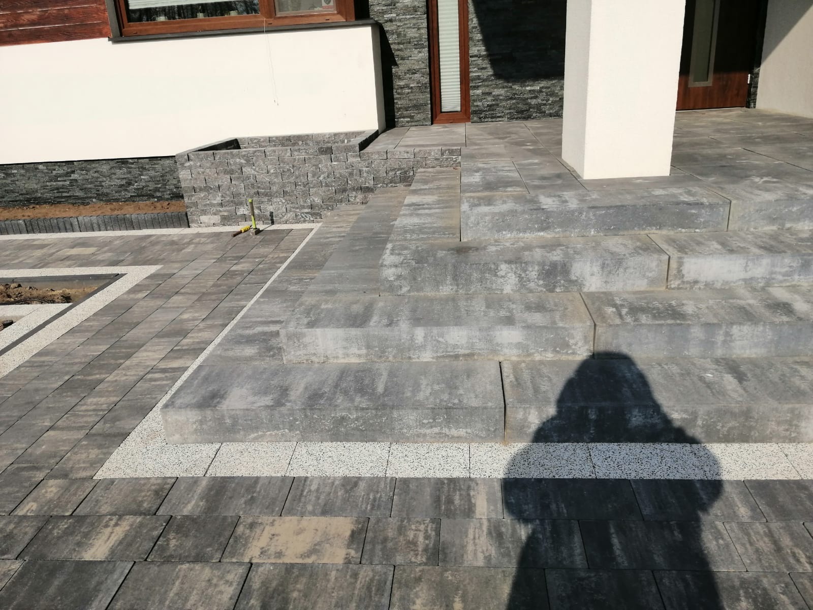 Schody wejściowe i chodnik wyłożone szarą kostką brukową z jasnymi akcentami, widoczny cień osoby fotografującej.