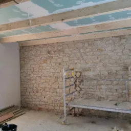 Wnętrze w trakcie remontu: surowa kamienna ściana, drewniane belki stropowe z fragmentami płyt gipsowo-kartonowych, metalowy podest roboczy, okno i materiały budowlane w nieuporządkowanym otoczeniu.