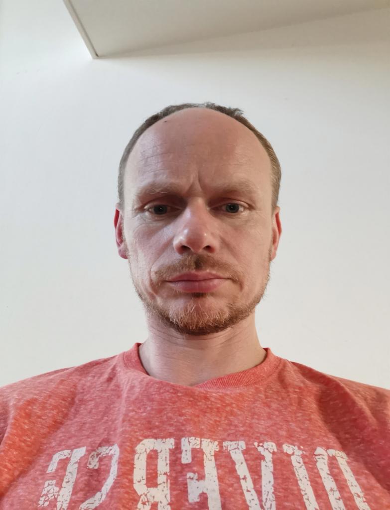 Portret mężczyzny z lekkim zarostem, ubranego w czerwoną koszulkę z białym napisem, na tle białej ściany i fragmentu sufitu. Ujęcie od dołu.