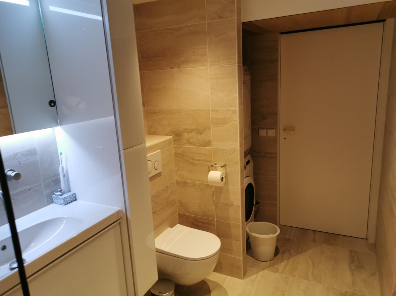 Nowoczesna łazienka z beżowymi płytkami imitującymi kamień, wisząca toaleta, umywalka z szafką w białym połysku, pralka ukryta za drzwiami.