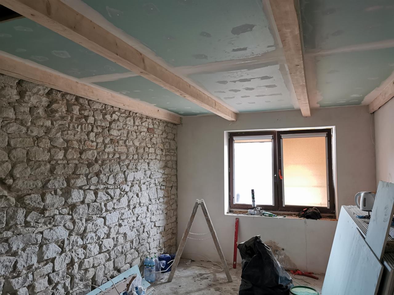 Wnętrze pokoju w Zduńskiej Woli w trakcie remontu: surowa kamienna ściana, drewniane belki pod sufitem z płytami gipsowo-kartonowymi, okno i drabina, ukazujące etap prac wykończeniowych.