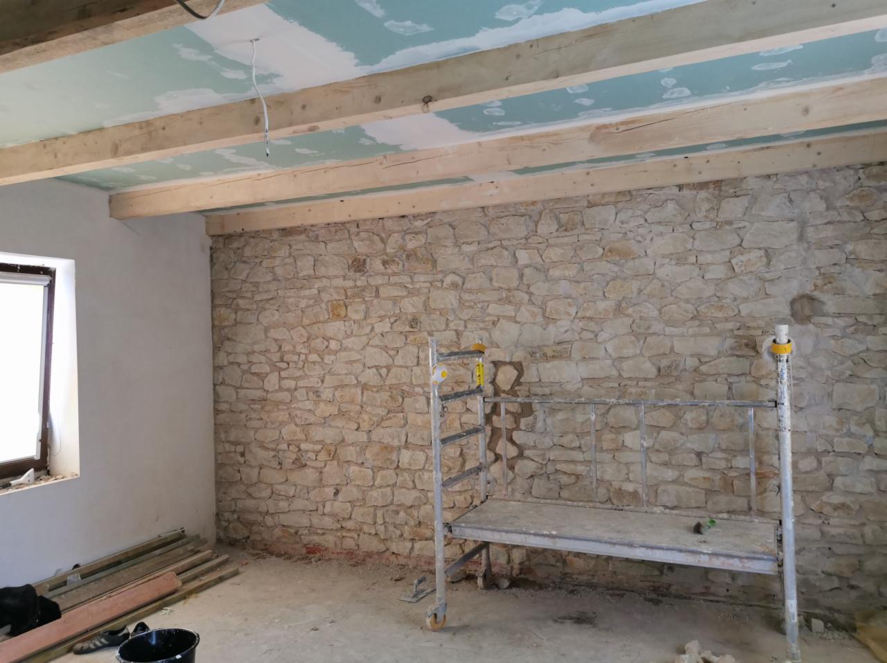 Wnętrze w trakcie remontu: surowa kamienna ściana, drewniane belki stropowe z fragmentami płyt gipsowo-kartonowych, metalowy podest roboczy, okno i materiały budowlane w nieuporządkowanym otoczeniu.