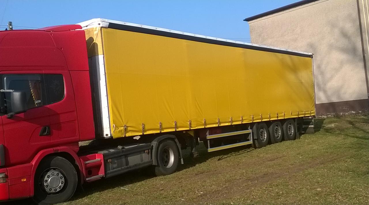 Czerwona ciężarówka z żółtą plandeką na trawiastym terenie, budynek w tle, błękitne niebo, widok z boku, trzy osie,  detal.