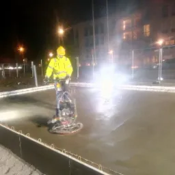 Pracownik w żółtym ubraniu ochronnym wyrównuje świeżo wylany beton za pomocą zacieraczki mechanicznej na budowie w nocy, widoczne oświetlenie placu budowy i budynek w tle.