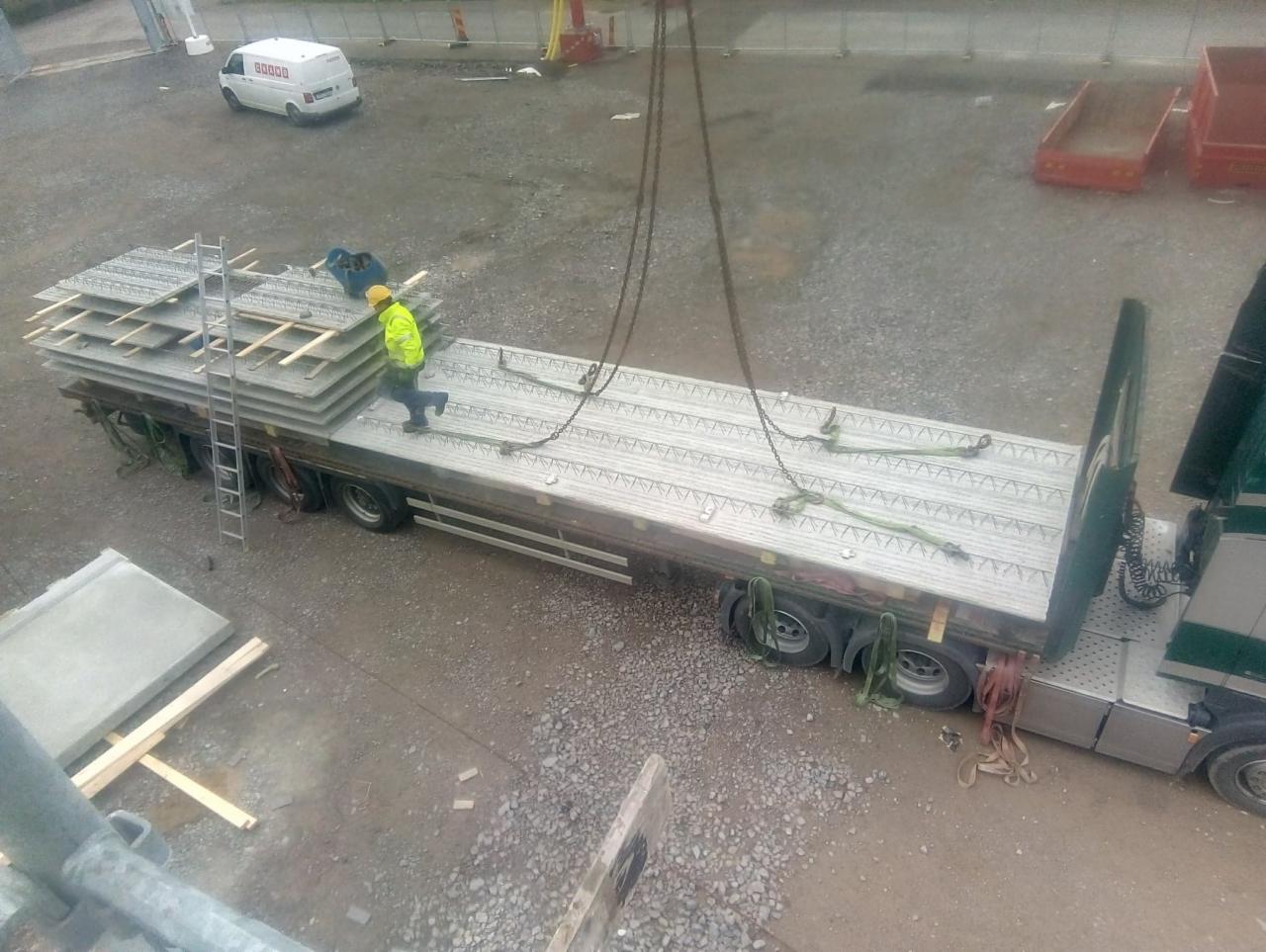 Widok z góry na pracownika w żółtej kurtce stojącego na naczepie ciężarówki załadowanej prefabrykowanymi elementami betonowymi, z dźwigiem w tle i białą furgonetką w oddali.