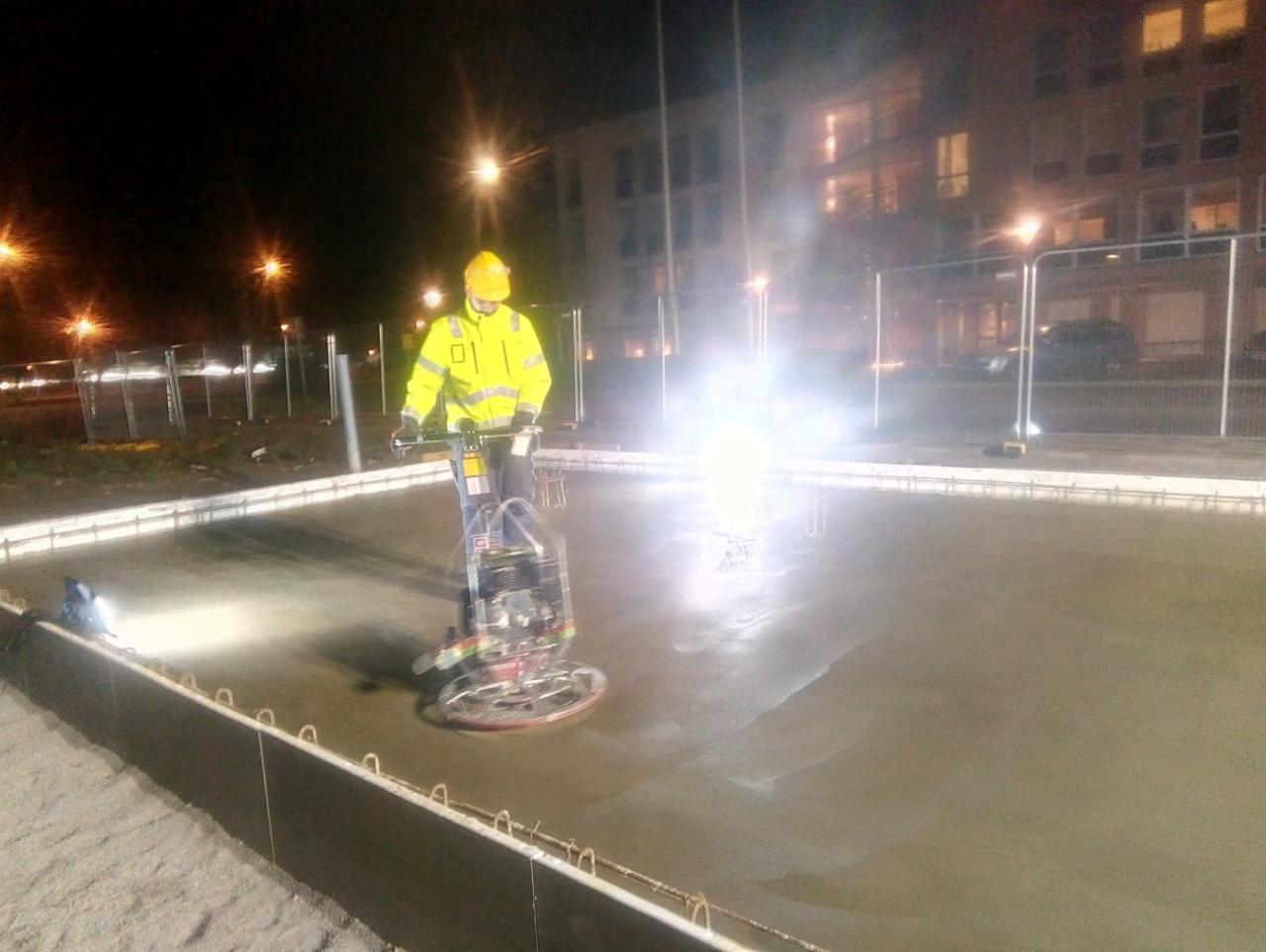 Pracownik w żółtym ubraniu ochronnym wyrównuje świeżo wylany beton za pomocą zacieraczki mechanicznej na budowie w nocy, widoczne oświetlenie placu budowy i budynek w tle.