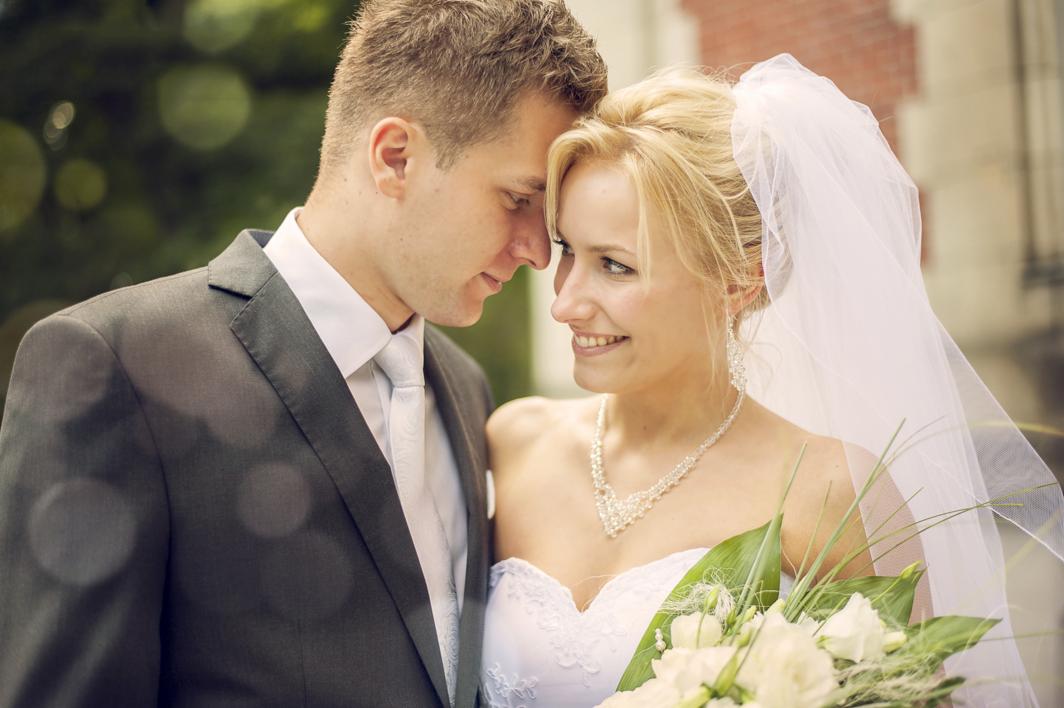 Uśmiechnięta panna młoda w białej sukni i z bukietem, opierająca głowę o pana młodego w szarym garniturze, na tle zieleni i ceglanego budynku.