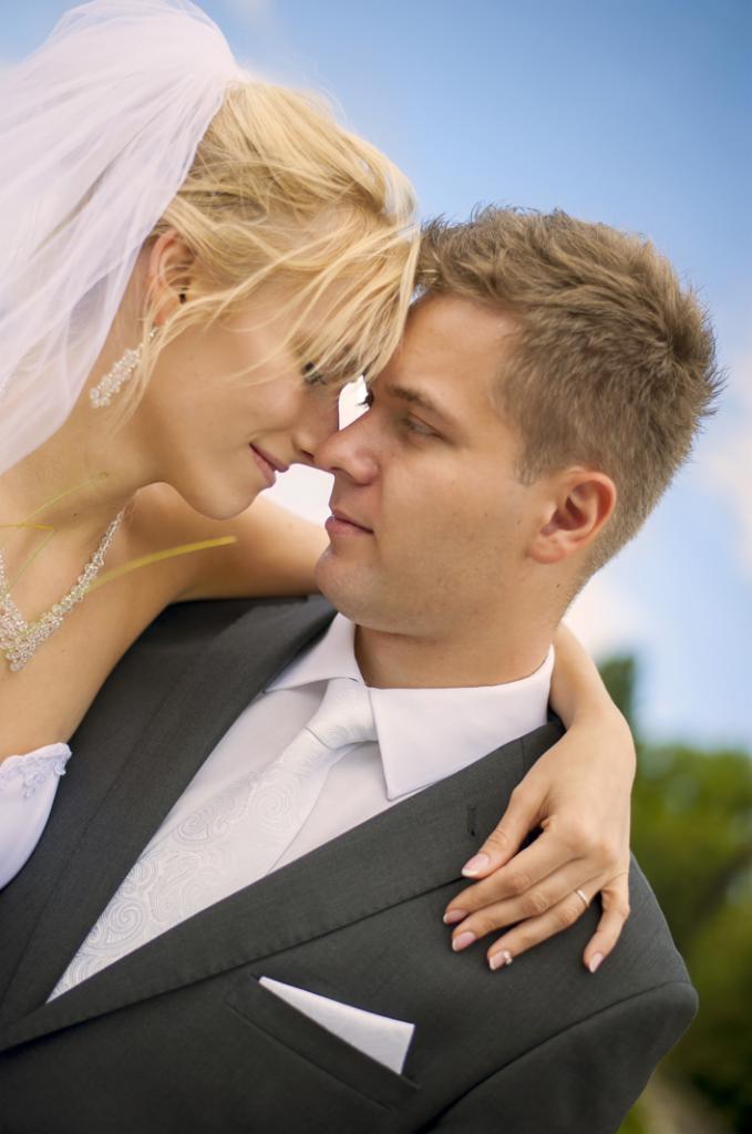 Panna młoda w welonie i z diamentową biżuterią, opiera głowę o pana młodego w szarym garniturze i białym krawacie w delikatny wzór, zbliżenie na twarze.