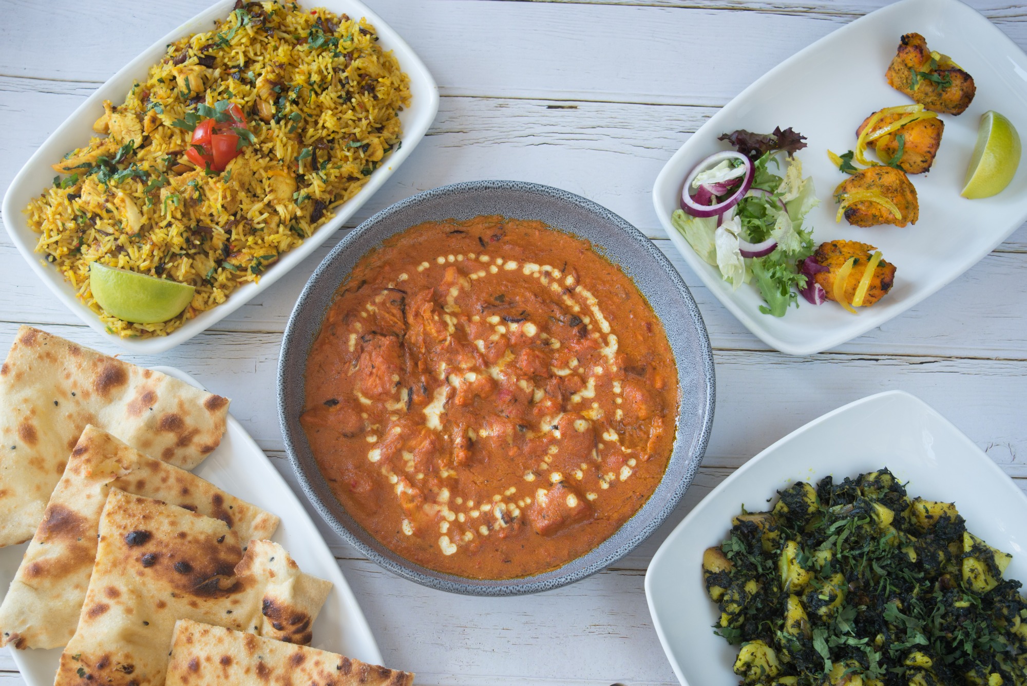 Widok z góry na stół z indyjskimi daniami: curry z kurczaka z kremowym wzorem, ryż biryani z kurczakiem i warzywami, pieczone placki naan, warzywa z ziemniakami i smażone kawałki kurczaka z cytryną.