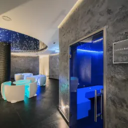 Nowoczesna aranżacja wnętrza łaźni z podświetlanymi meblami, ciemną podłogą i ścianami o fakturze betonu, oświetleniem imitującym gwiazdy na suficie oraz tabliczką informacyjną 'Łaźnia Bałtycka'.