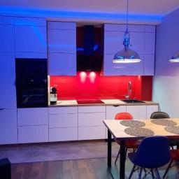 Meb-Le - Nowoczesna kuchnia z białymi szafkami, czerwonym panelem i drewnianym blatem. Nad stołem wiszą lampy w stylu industrialnym, a całość oświetlona jest niebieskim światłem.