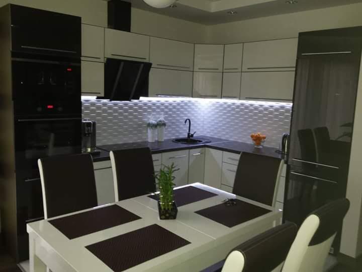 Nowoczesna kuchnia z białymi szafkami i czarnymi akcentami, oświetleniem LED pod szafkami wiszącymi, stół z czterema krzesłami na pierwszym planie, roślina doniczkowa na stole.