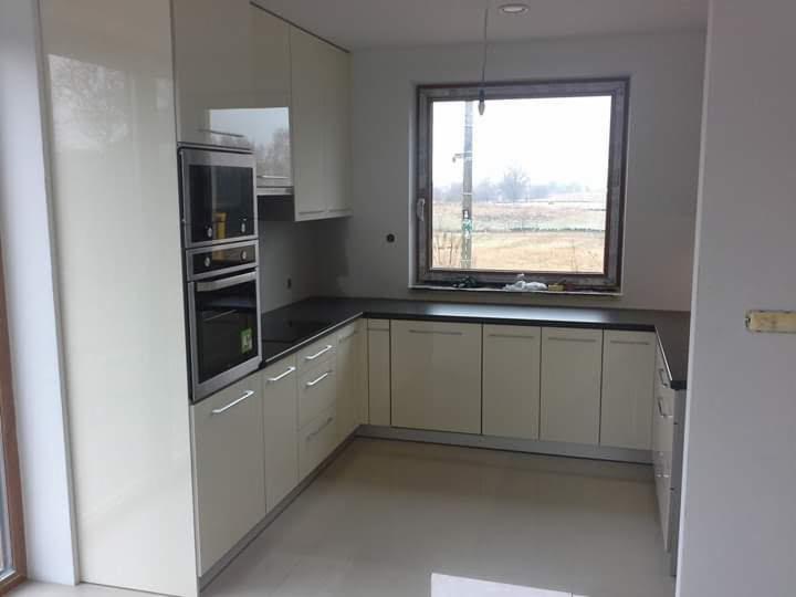 Jasna kuchnia z kremowymi, lakierowanymi frontami szafek, czarnym blatem i wbudowanym piekarnikiem, widok na pole za oknem.