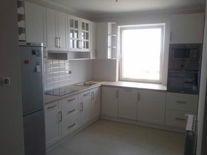 Jasna, funkcjonalna kuchnia z białymi frontami szafek, płytą indukcyjną i wbudowanym piekarnikiem, oświetlona naturalnym światłem z okna.