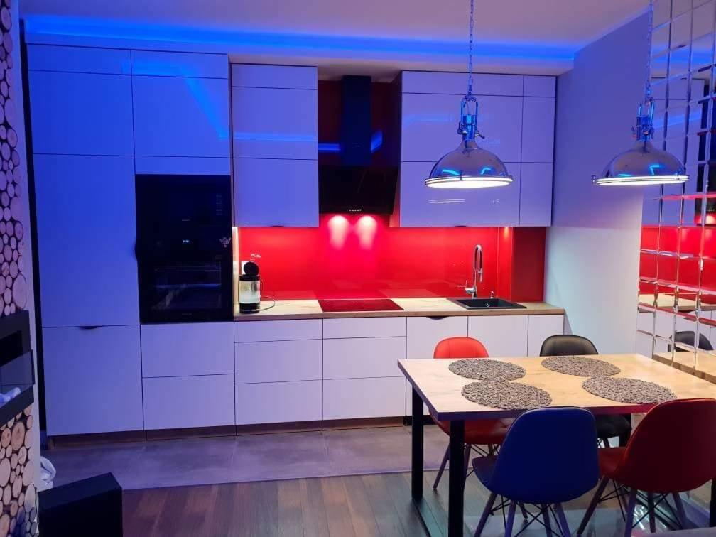 Nowoczesna kuchnia z białymi szafkami, czerwonym panelem i drewnianym blatem. Nad stołem wiszą lampy w stylu industrialnym, a całość oświetlona jest niebieskim światłem.