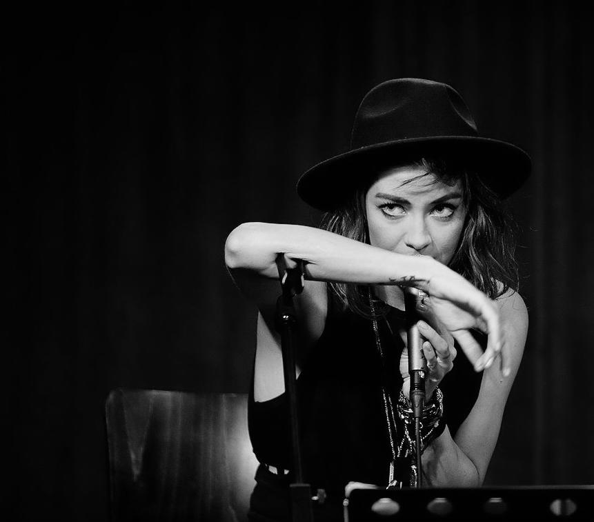 Czarno-białe zdjęcie piosenkarki w kapeluszu, trzymającej mikrofon, z tatuażem na przedramieniu i intensywnym spojrzeniem w obiektyw.