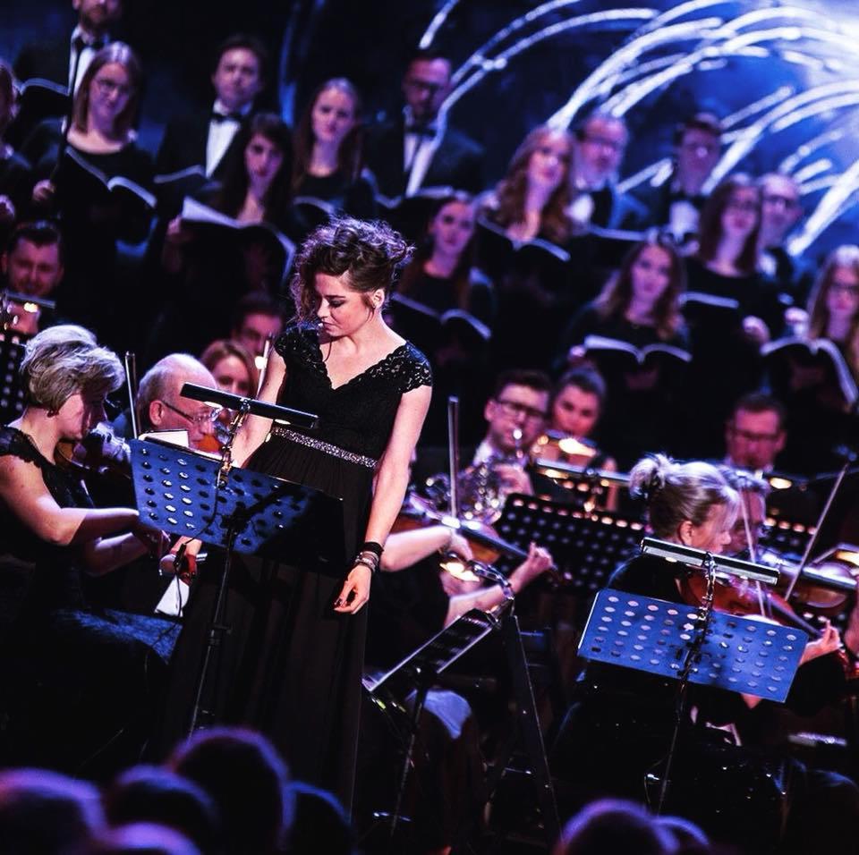 Wokalistka w czarnej sukni na scenie przed chórem i orkiestrą smyczkową podczas koncertu.