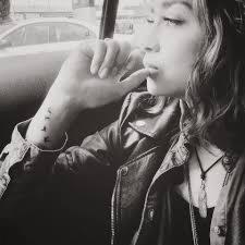 Czarno-białe portretowe ujęcie kobiety w skórzanej kurtce, siedzącej w samochodzie, z tatuażem trzech trójkątów na nadgarstku i zamyśloną miną.