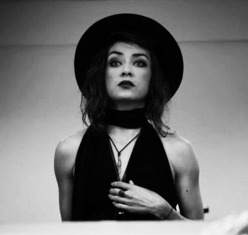 Czarno-białe ujęcie kobiety w kapeluszu i czarnej sukience, z wyrazistym makijażem, sugerujące artystyczny charakter występu lub sesji fotograficznej.