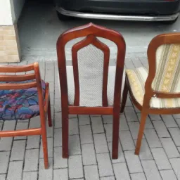 Trzy krzesła przed garażem: odnowione krzesło z motywem kwiatowym, krzesło z wysokim oparciem i jasną tapicerką oraz krzesło z beżowo-złotymi paskami.