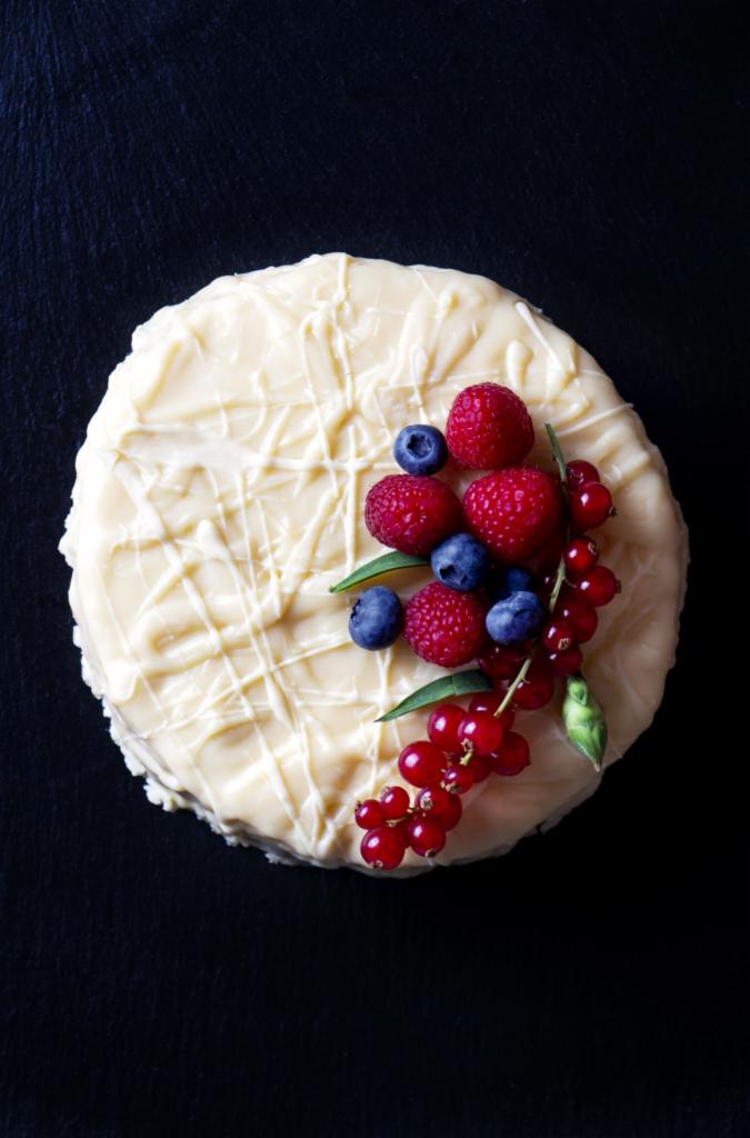 Tort z białej czekolady udekorowany malinami, borówkami i czerwoną porzeczką na czarnym tle, widok z góry.