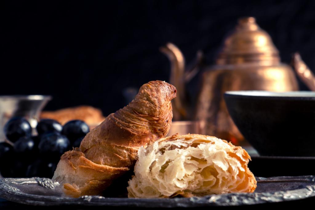 Kulinarna martwa natura w Warszawie: przekrojony croissant na srebrnej tacy, obok kiść ciemnych winogron, w tle mosiężny imbryk i czarka na herbatę.
